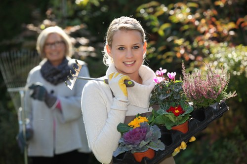 A young woman with light-colored hair tied back, wearing a white sweater and gardening gloves, is standing outdoors in a garden during daylight, holding a black tray filled with various flowering plants and foliage in shades of pink, purple, yellow, and green. Behind her, slightly out of focus, an older woman wearing glasses and a grey scarf over a white jacket is also engaged in gardening, holding a gardening tool. The garden features a well-maintained lawn with dense, colorful shrubbery and trees in the background, indicating a sunny, temperate day with natural light illuminating the scene. The setting appears to be a landscaped outdoor space suitable for gardening services, with healthy grass, vibrant plants, and a tidy environment, aligning with professional gardening and lawn care activities in Canary Wharf or nearby areas.