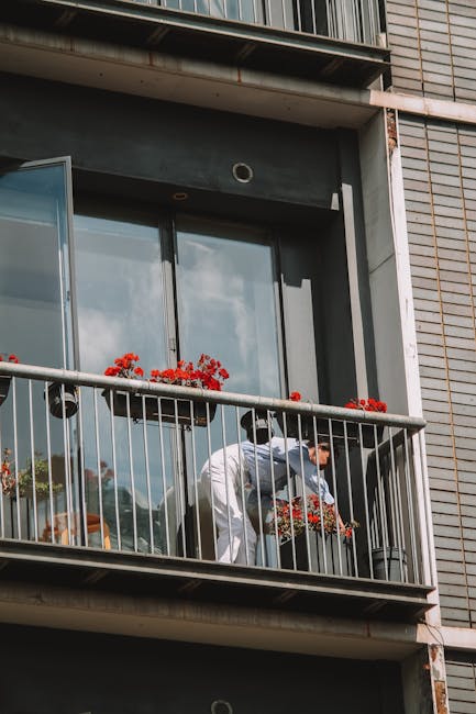 A person dressed in white, bending slightly forward on a balcony of a modern multi-story building, tending to potted red flowering plants placed along the metal railing. The balcony has glass doors wi