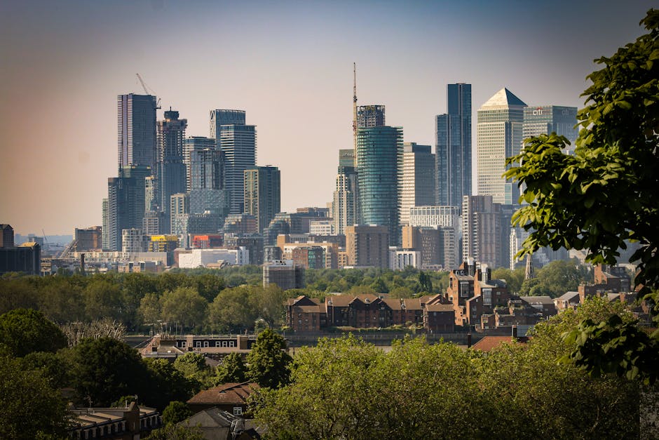 A panoramic cityscape featuring a dense cluster of modern skyscrapers and high-rise buildings, with reflective glass facades capturing the sky. In the foreground, there is a lush green park with tight
